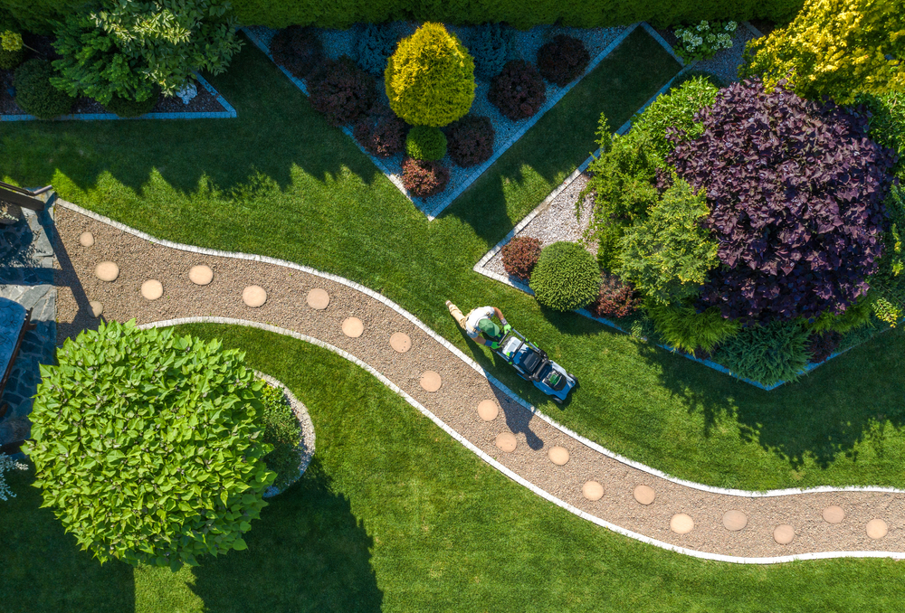 overhead shot of garden mowing bright green grass along winding path in landscaped, sunny garden with sectioned trees.