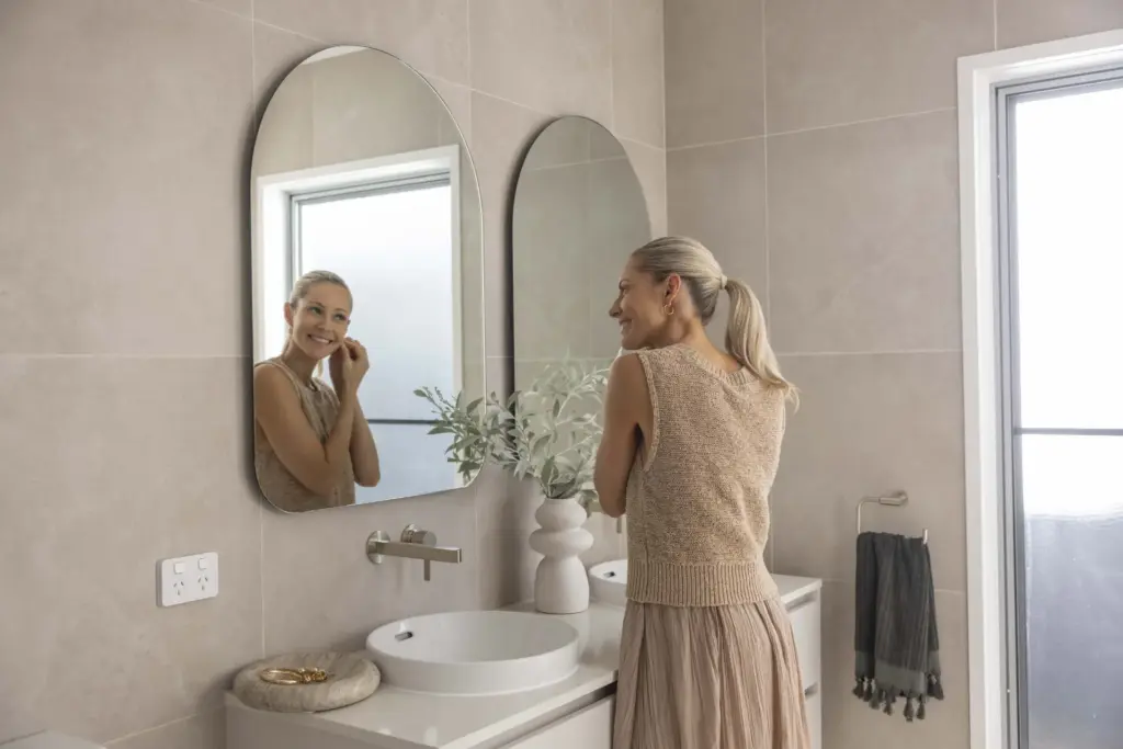 A woman with blond hair in a ponytail smiles while putting on an earring, standing in front of a mirror in a modern bathroom with light-colored tiles and a white sink.