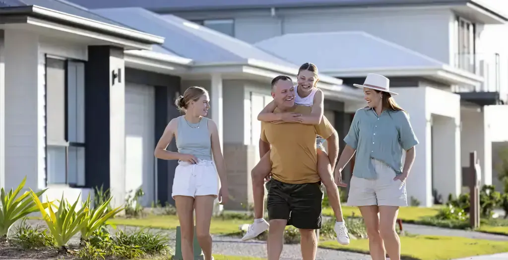 A group of four people, two adults and two children, walk and laugh together on a sidewalk in a modern suburban neighborhood with white houses and greenery on a sunny day.