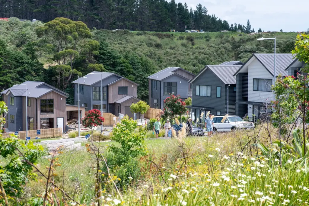 A group of people gathers near parked cars on a suburban street lined with modern homes and greenery, with trees and hills in the background under a partly cloudy sky.