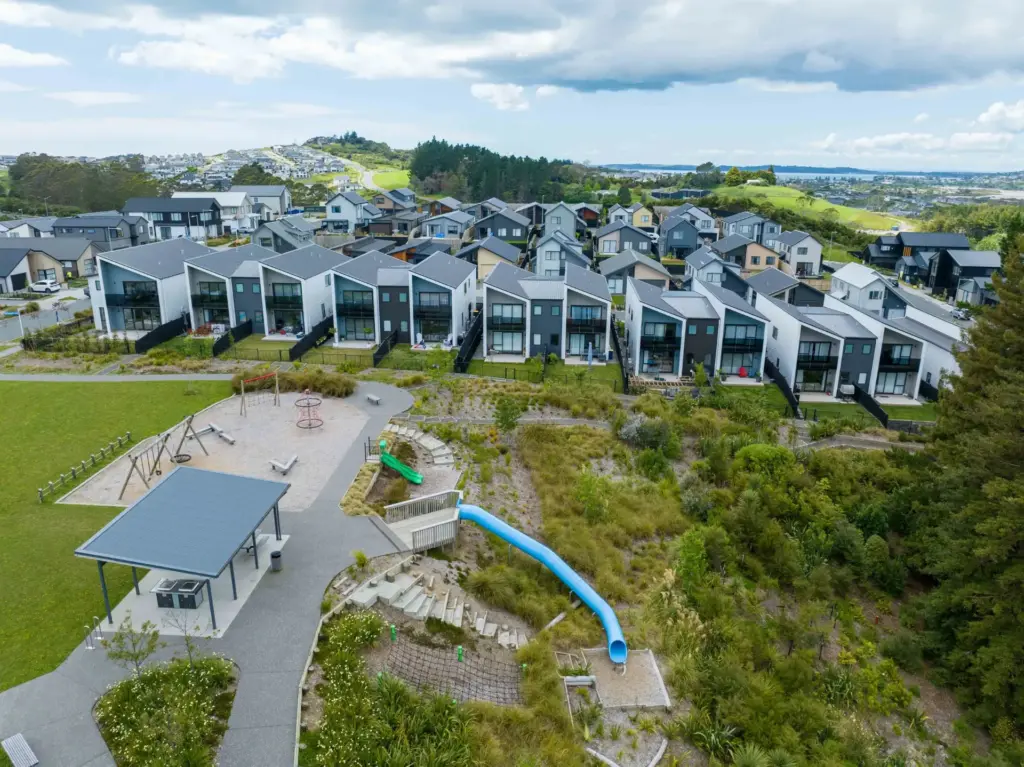 A suburban neighborhood with rows of modern houses facing a park featuring a playground, a blue slide, a sheltered picnic area, and landscaped greenery, with hills and more houses in the background.