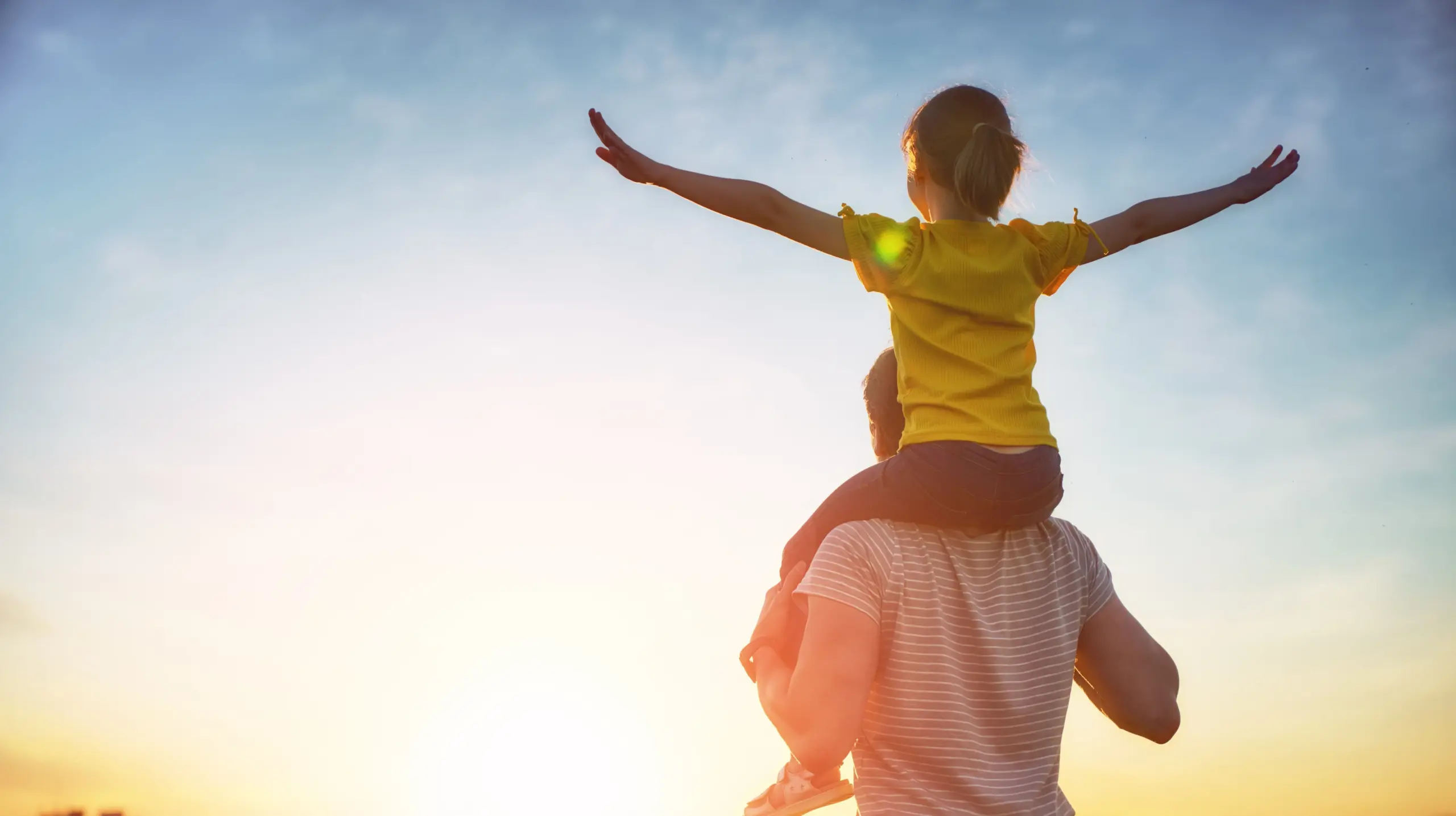 A child wearing a yellow shirt sits on an adult's shoulders with arms outstretched, facing a bright sunset or sunrise sky. The scene conveys joy and togetherness.