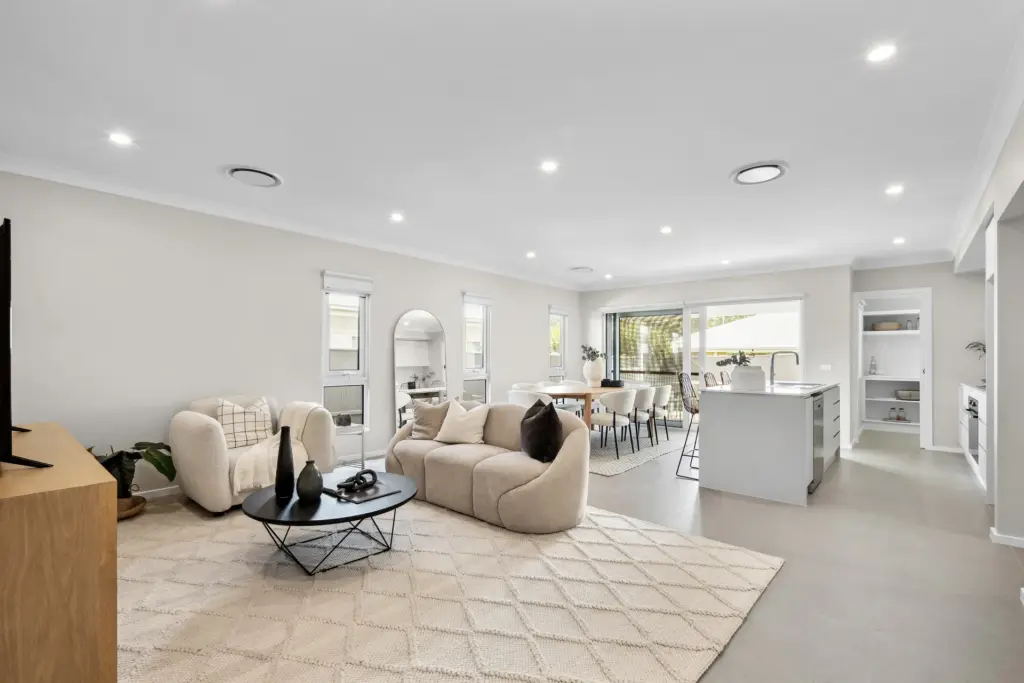 Modern open-plan living space featuring a beige sofa and armchair, round coffee table, dining area, and kitchen with island. Large windows and glass doors provide natural light. Neutral color palette and minimalist decor.