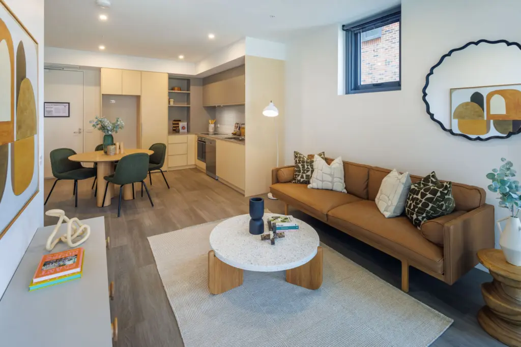 Modern apartment living room and kitchen with a brown sofa, round coffee table, dining area with green chairs, wall art, and neutral decor. Light wood and beige tones create a bright, inviting space.