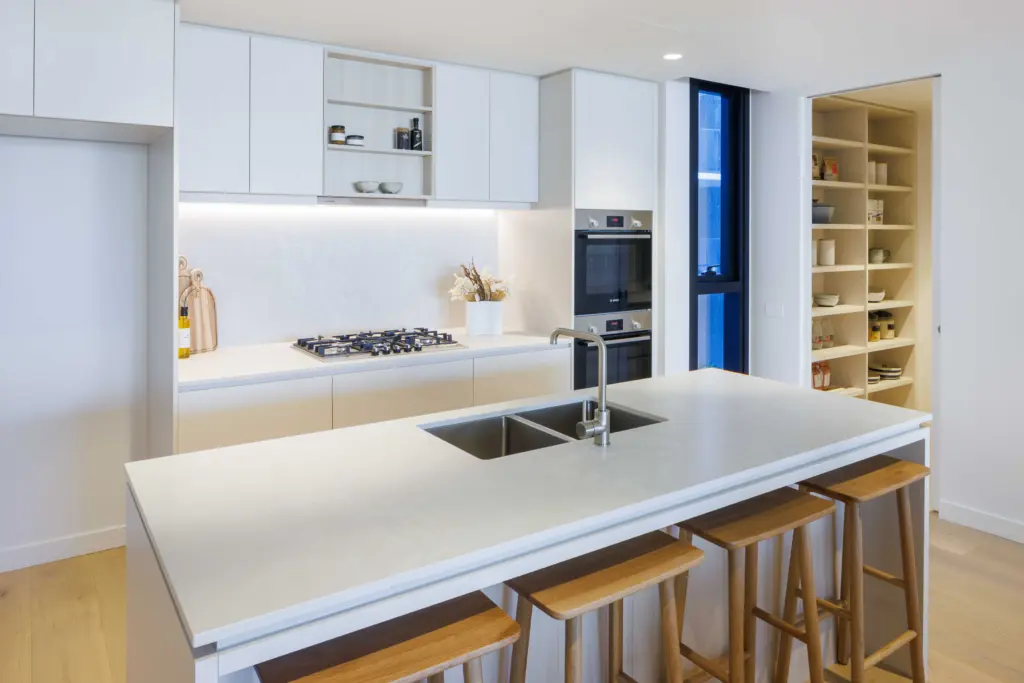 Modern kitchen with a large white island featuring a built-in sink and four wooden stools. Open shelving, gas stove, oven, and a walk-in pantry filled with food items are visible in the background.