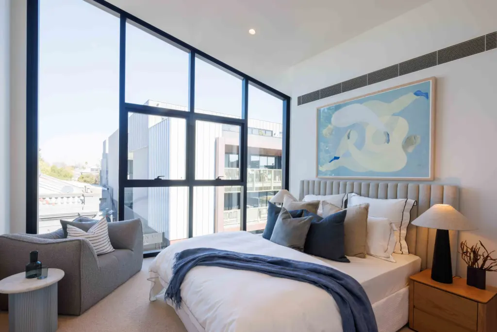 Modern bedroom with large floor-to-ceiling windows, a double bed with blue and gray pillows, a blue throw, a gray armchair, bedside tables with lamps, and abstract artwork above the bed.