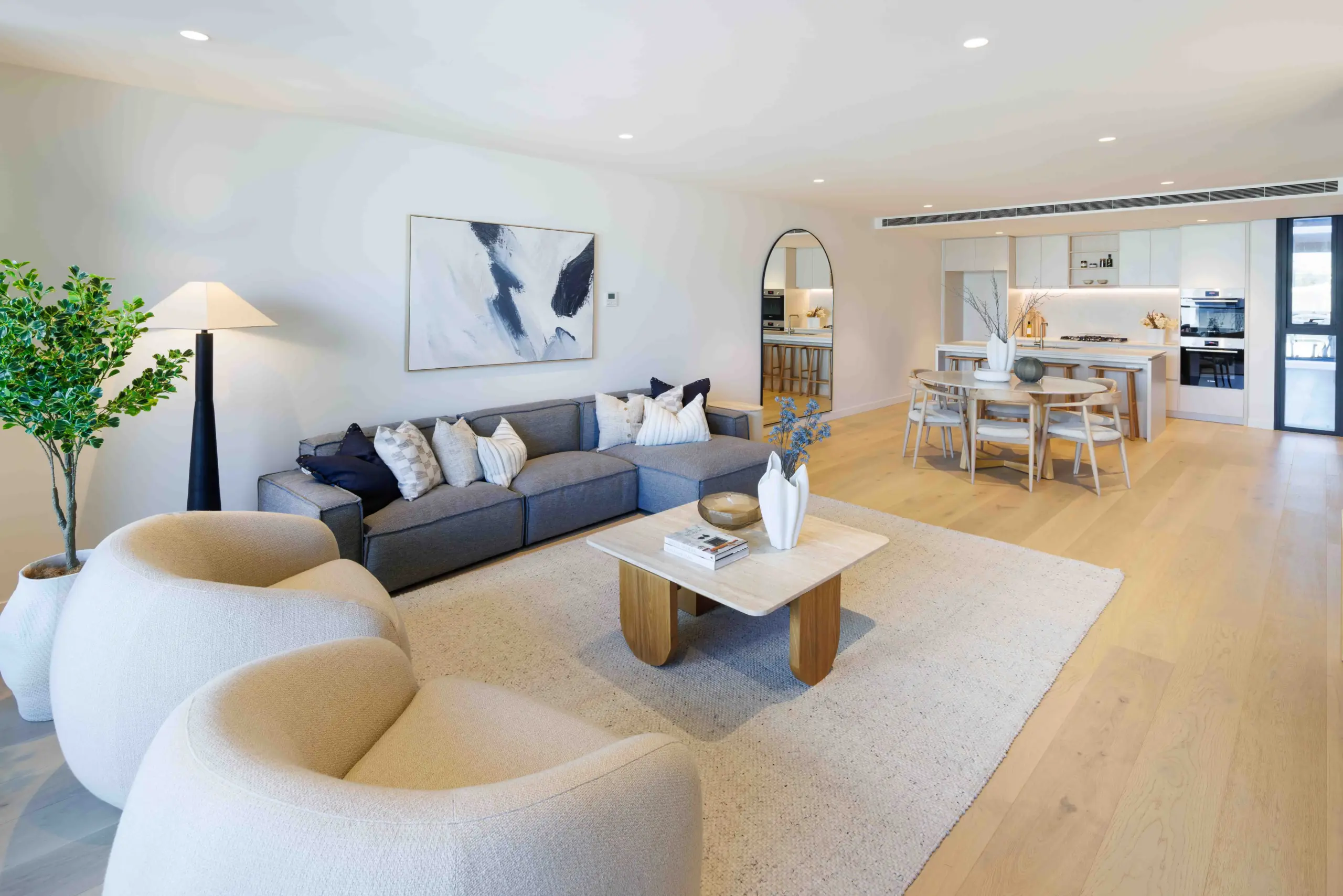 A modern, open-concept living room and kitchen with light wood floors, a gray sofa, two cream chairs, a light rug, a wood coffee table, dining table with chairs, and minimalist décor.