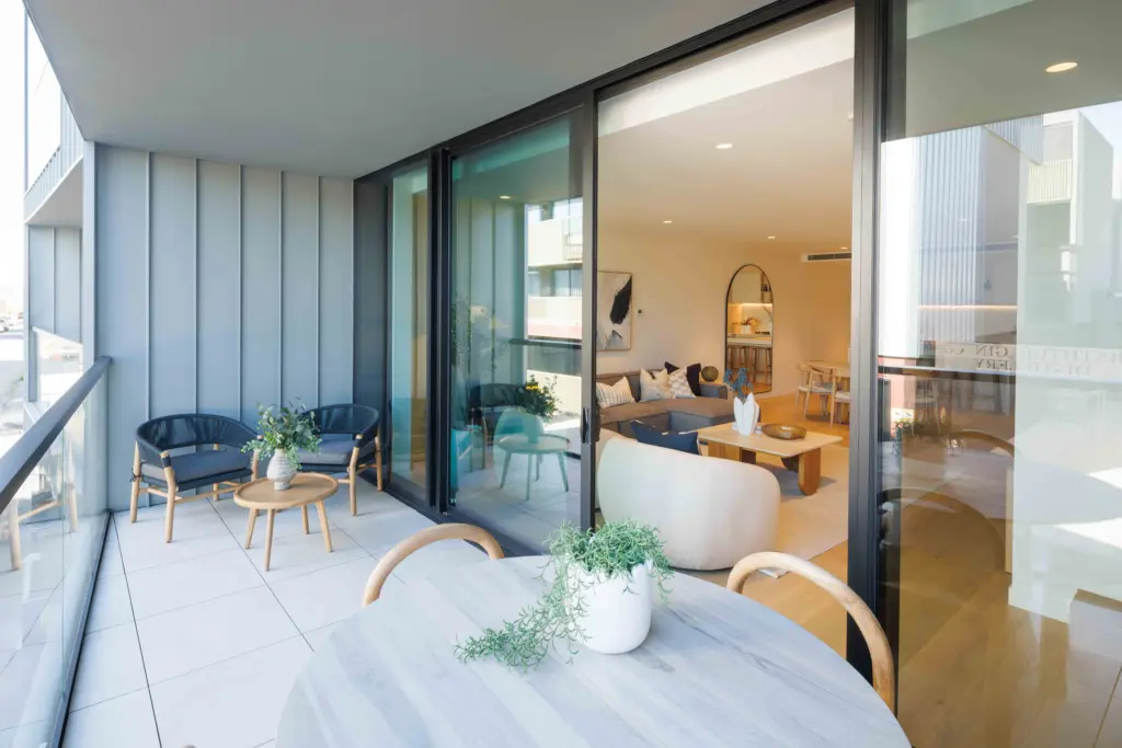 Modern balcony with a round table, potted plant, and chairs, overlooking a stylish living room through large glass sliding doors. The living room has cozy seating, a coffee table, and contemporary decor.