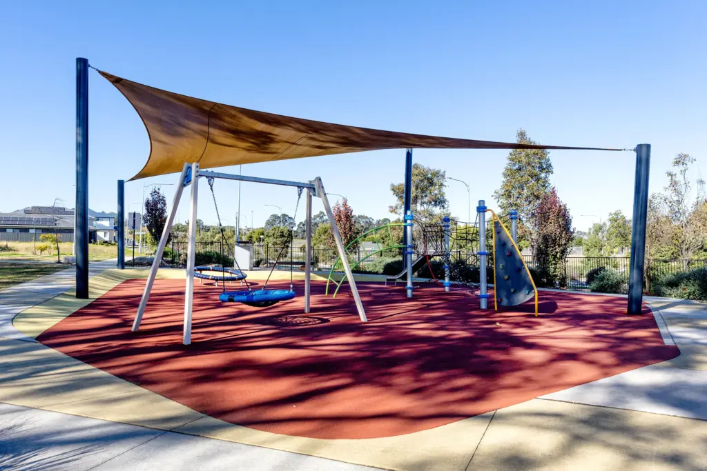 A modern playground with swings and climbing equipment on a red rubber surface, covered by a shade sail, surrounded by trees and a clear blue sky.