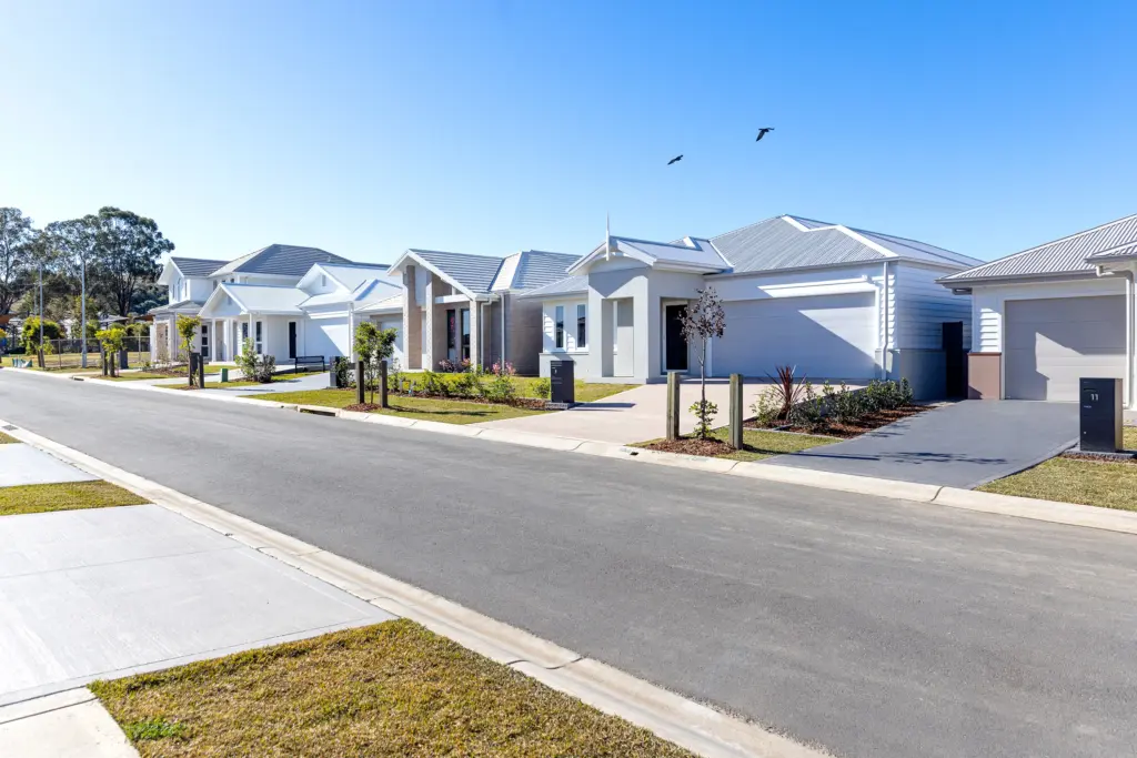 A quiet suburban street lined with modern, single-story houses, neatly manicured lawns, clear blue sky, and two birds flying overhead.