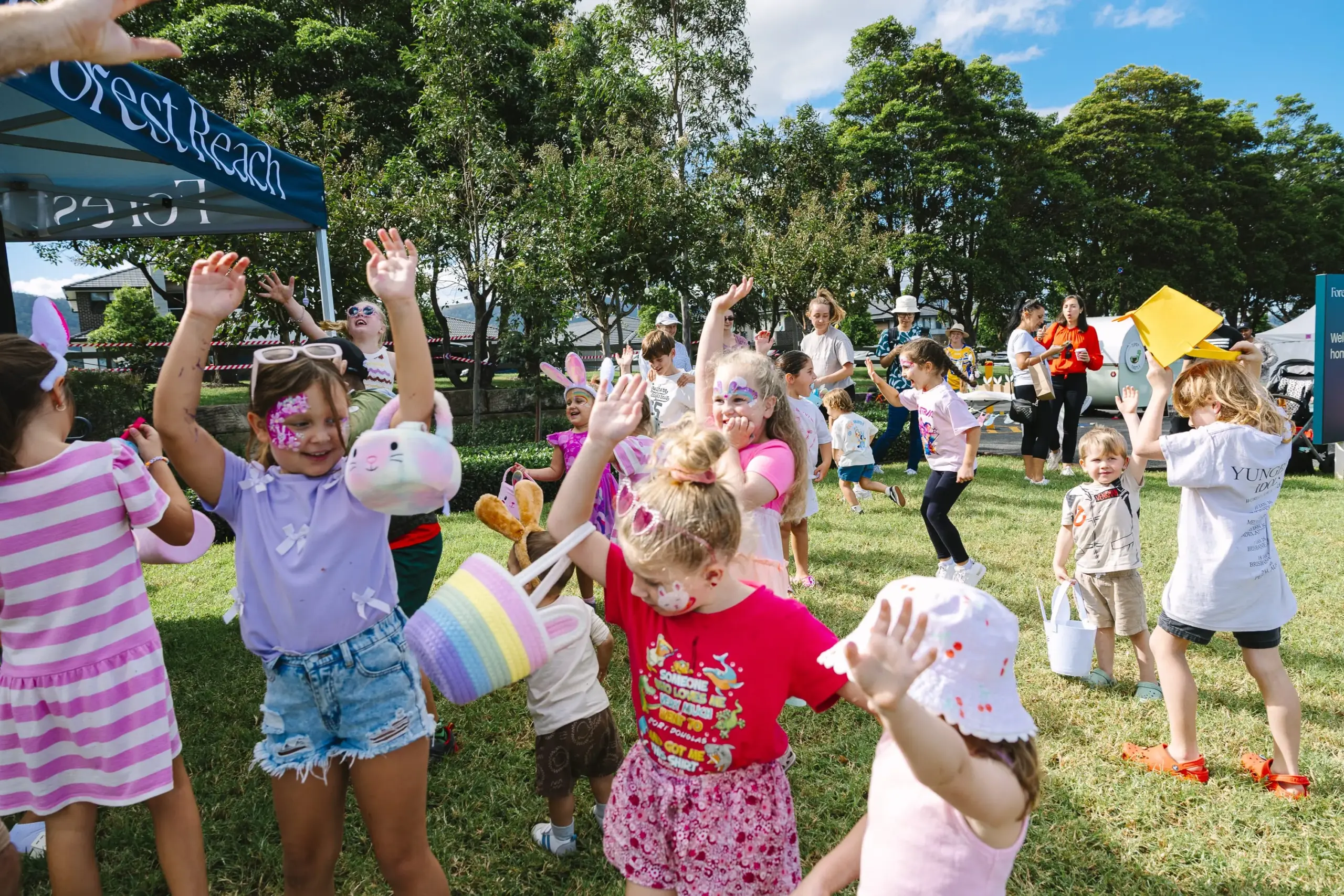 Children celebrate outdoors at a community event, some with painted faces, baskets, and bunny ears. Adults and kids are gathered on green grass under a sunny sky with trees and tents in the background.