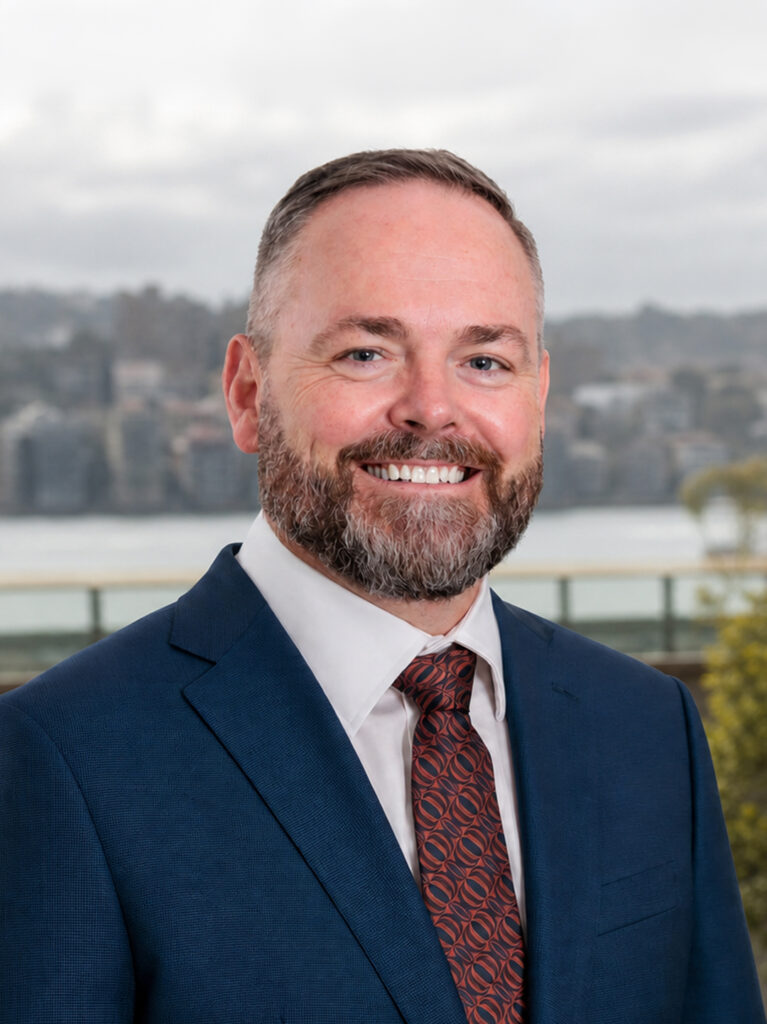 A man in a blue suit and patterned tie smiles outdoors, with a blurred background of water and buildings on a cloudy day.