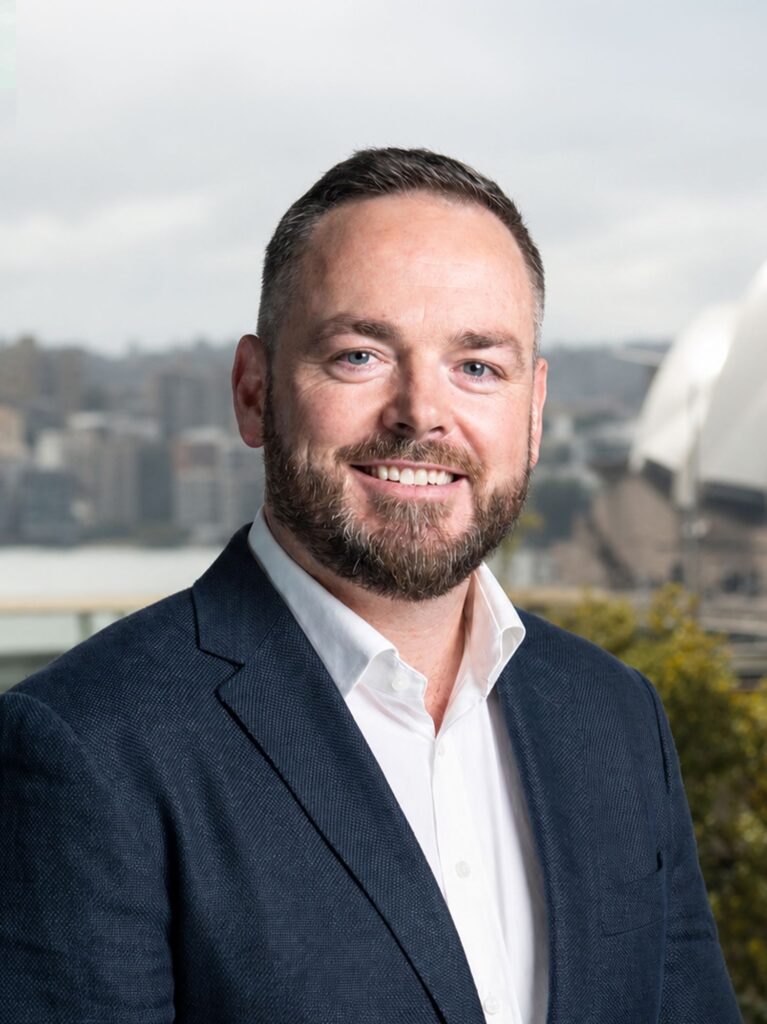 A bearded man wearing a navy blazer and white shirt smiles in front of a cityscape with the Sydney Opera House in the background.