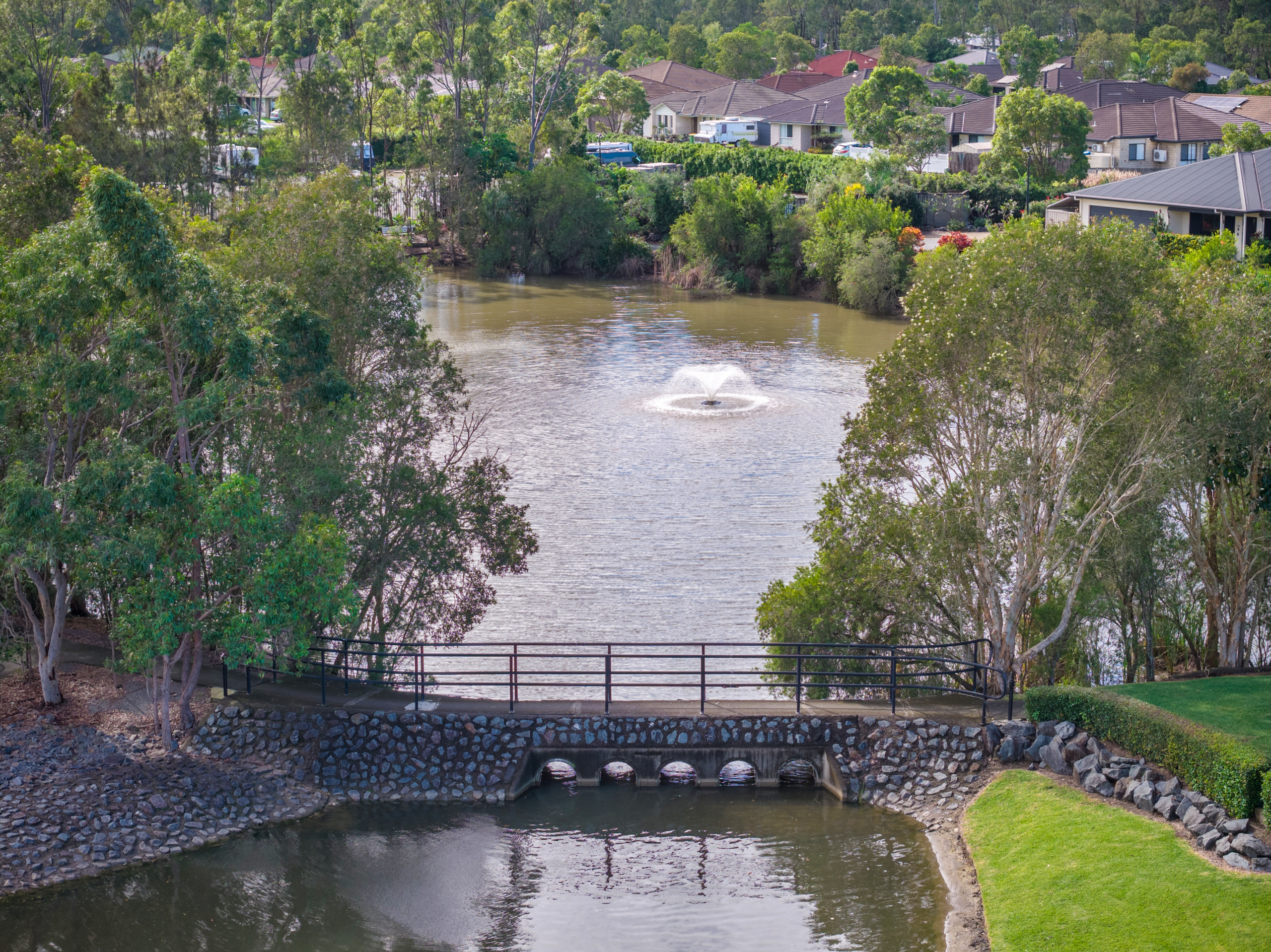 A small river with a water fountain in the center, surrounded by trees. A wooden footbridge crosses a narrow section, and houses are visible in the background under a cloudy sky.