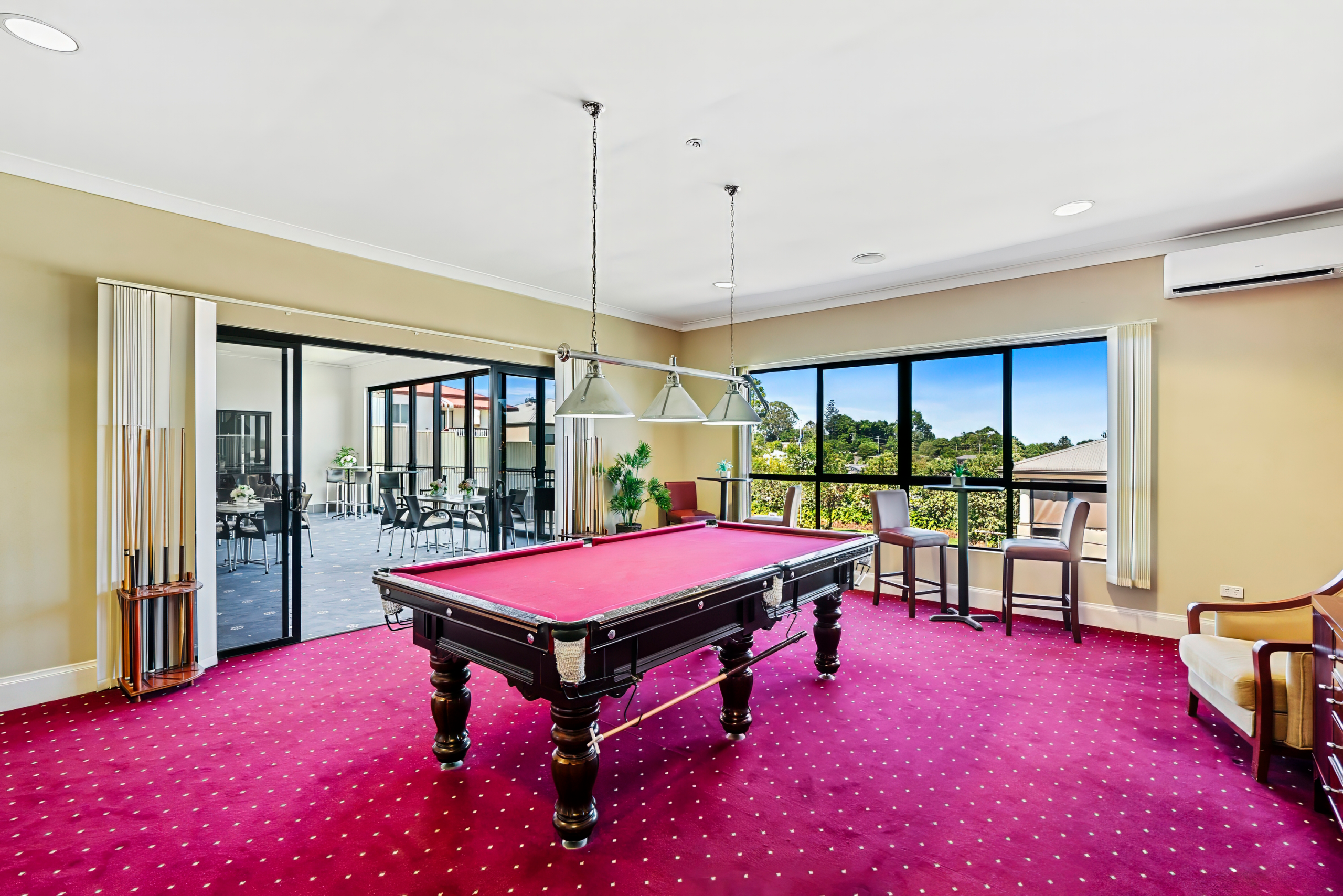 A room with a red pool table on red carpet, cue rack, chairs, windows with outdoor view, and glass doors leading to a sunroom with more seating.